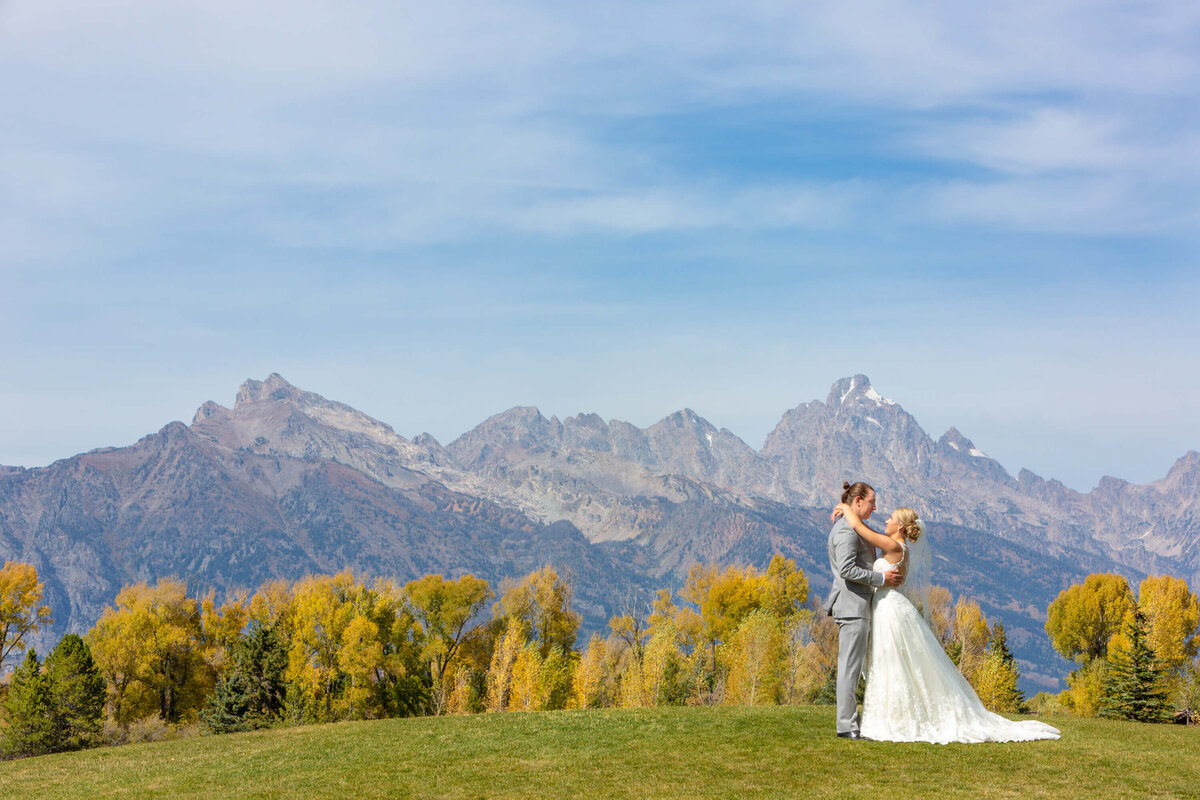 A bride and groom share a romantic moment on a grassy ridge at Jackson Hole Golf and Tennis Club with the Grand Tetons behind them. The golden fall foliage enhances the scenic wedding setting.