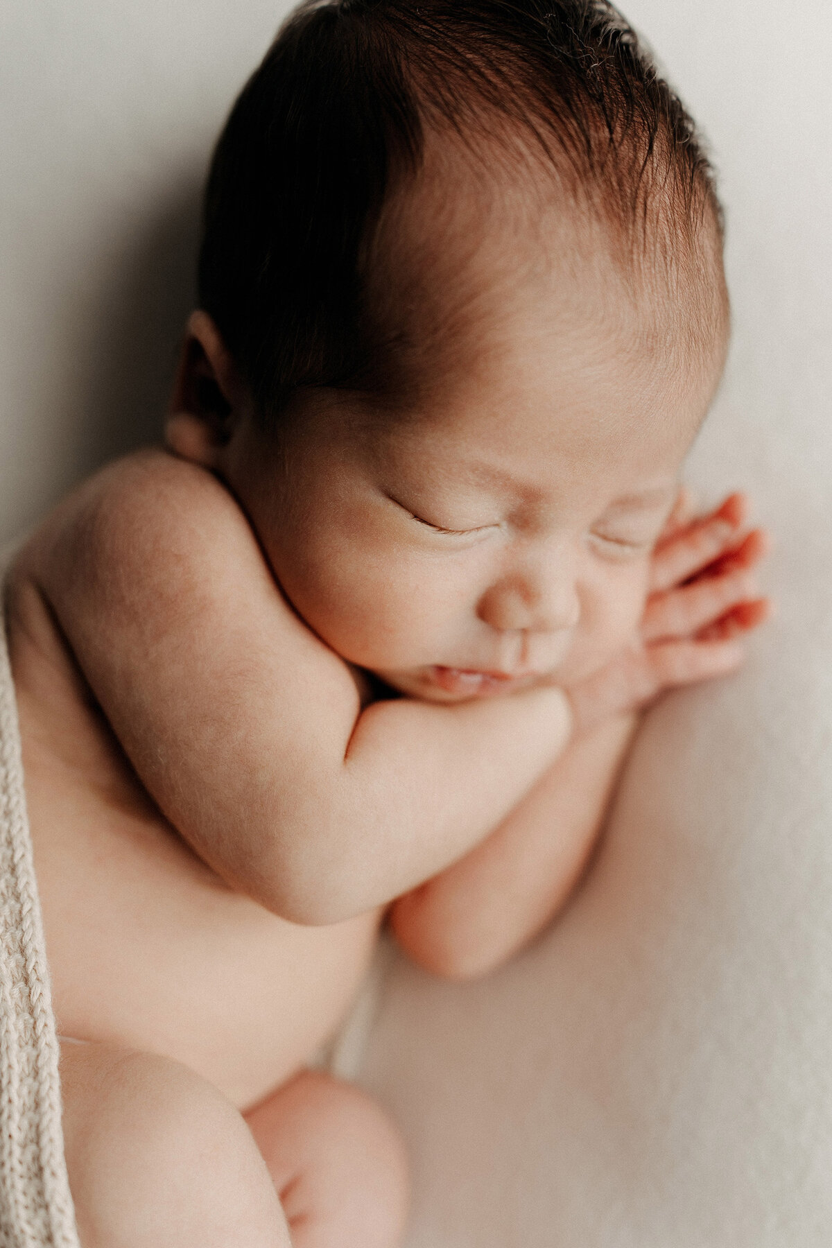 Newborn baby sleeping peacefully on their side, with hands tucked near their face and wrapped loosely in a soft knit blanket.