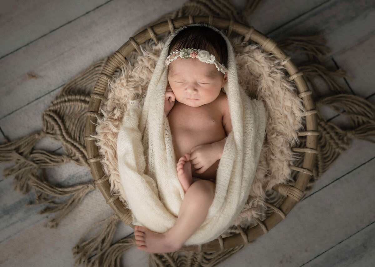 A newborn baby girl sleeps in a woven basket on a fur blanket in a studio