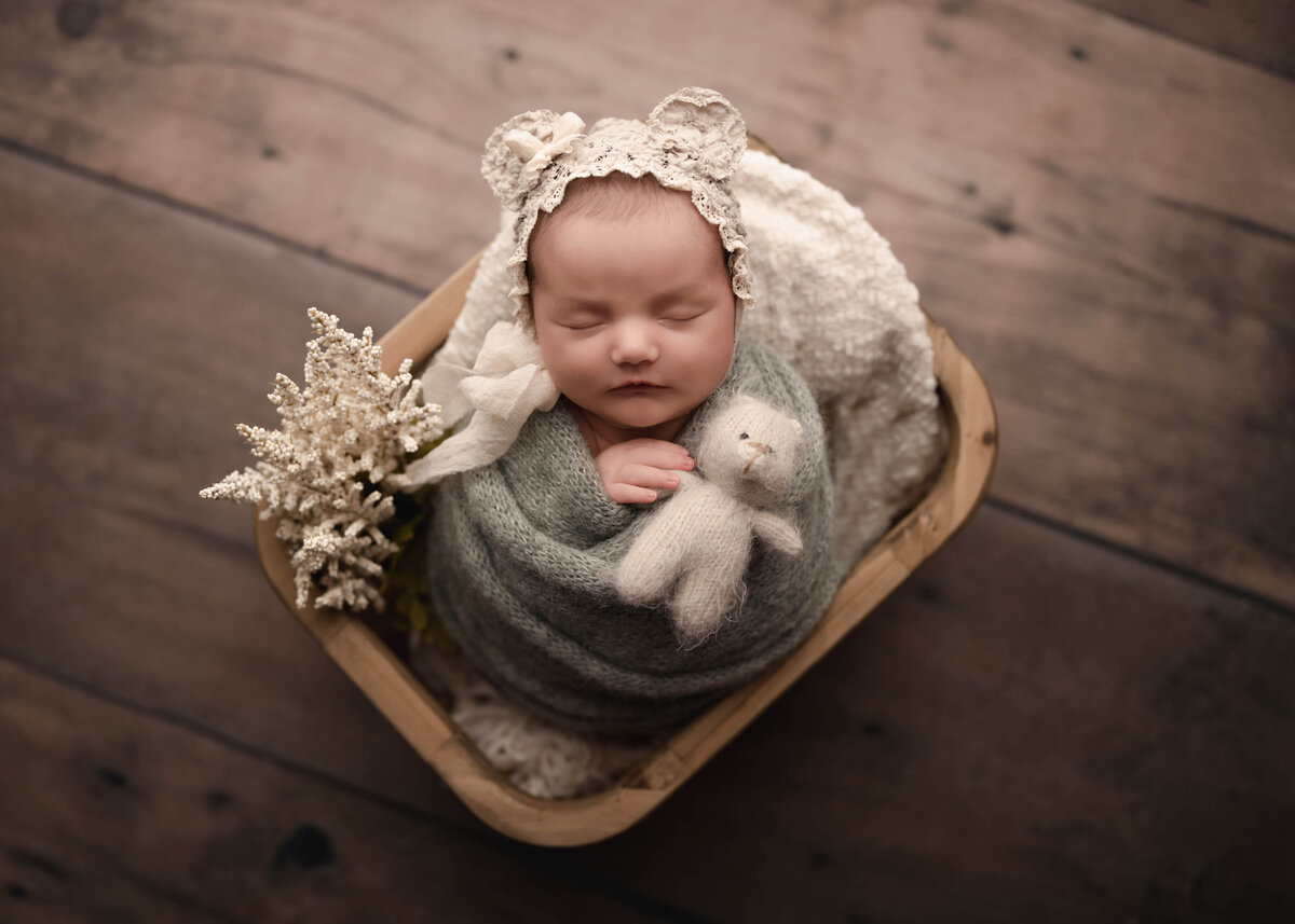 Aerial image captured at Corona newborn photoshoot. Baby girl swaddled in a sage green knit wrap and lace bonnet with teddy bear ear embellishments. Her fingers are peeking out of the wrap and touching a small felt teddy bear. Captured by best Corona newborn photographer Bonny Lynn Photography