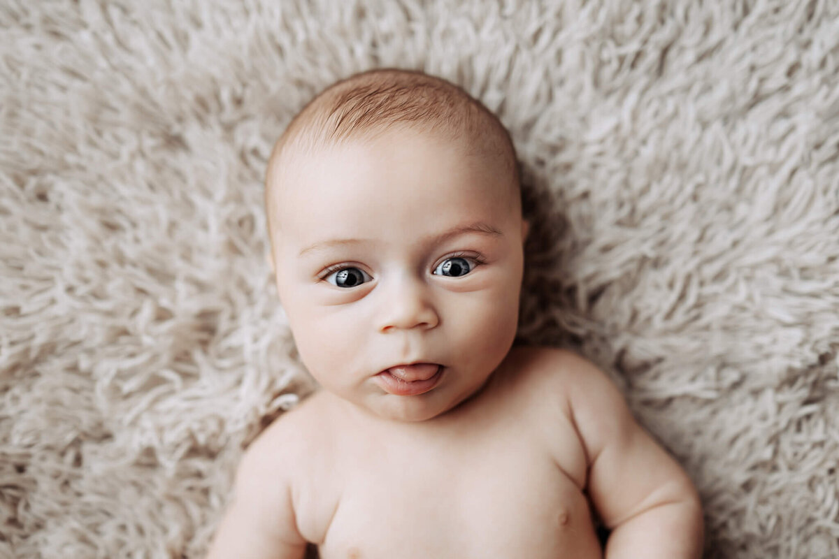 A baby with wide eyes and a curious expression lies on a soft, fluffy beige blanket. Captured by a portrait photographer in Bloomington, Illinois, the baby's slight smile and direct gaze create a warm, cozy atmosphere ideal for family photography.