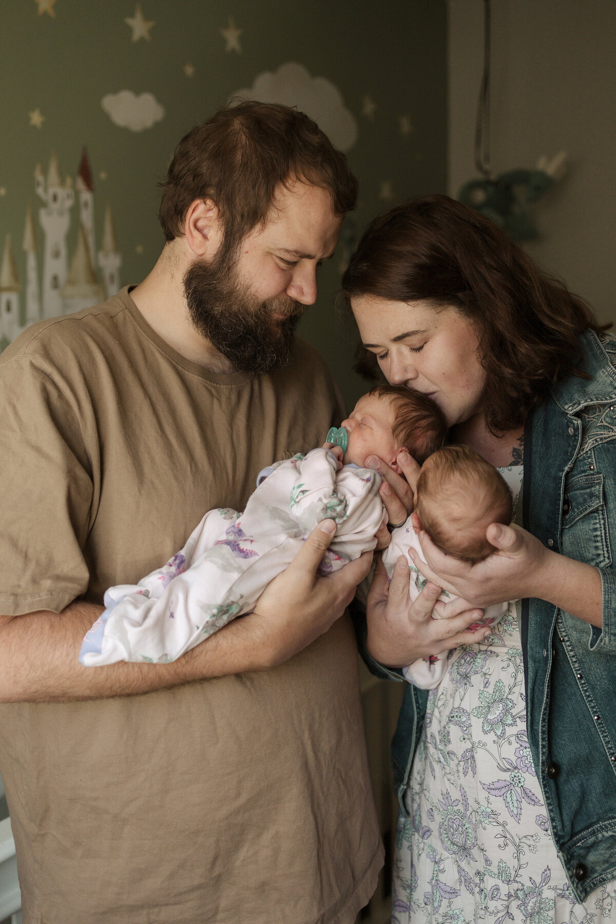 mom kissing top of newborn twins head while holding the other twin newborn with dad looking down at mom and baby twin girls with binkies in their mouths during in home newborn session in firestone colorado