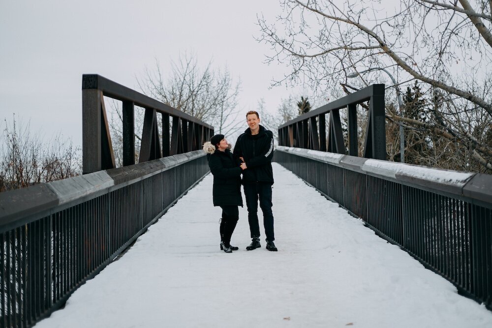 An Edmonton couple embrace on a bridge covered in snow in their black coats