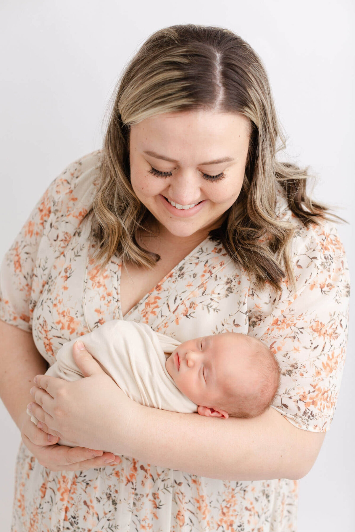 Woman with blonde hair and wearing a beige dress with rust colored flowers, she has professional hair and makeup done. She is smiling down at her newborn baby whom she is holding in her arms. Baby is sleeping peacefully in Mama's arms.
