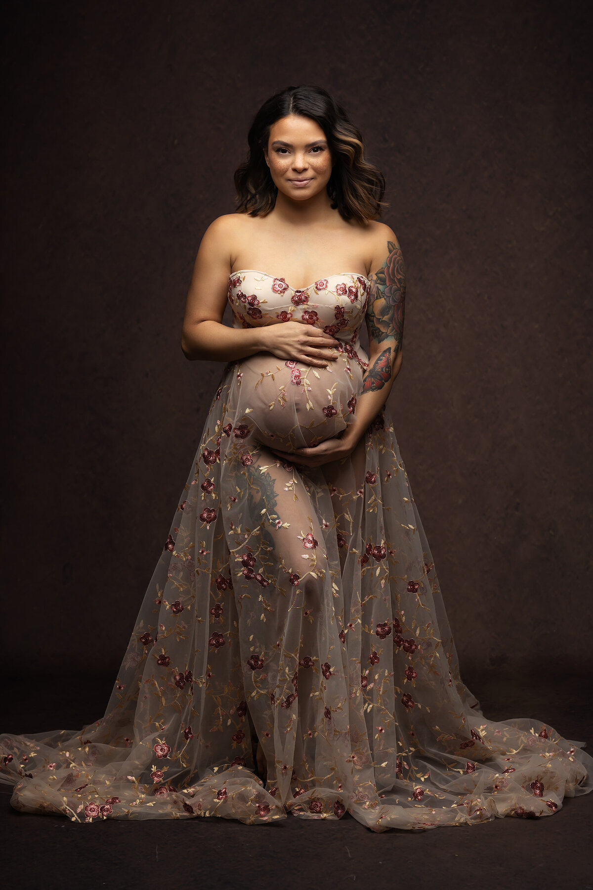 Pregnant woman in a strapless gown with red floral embroidery, gently cradling her belly. She stands against a dark background, radiating calm confidence.