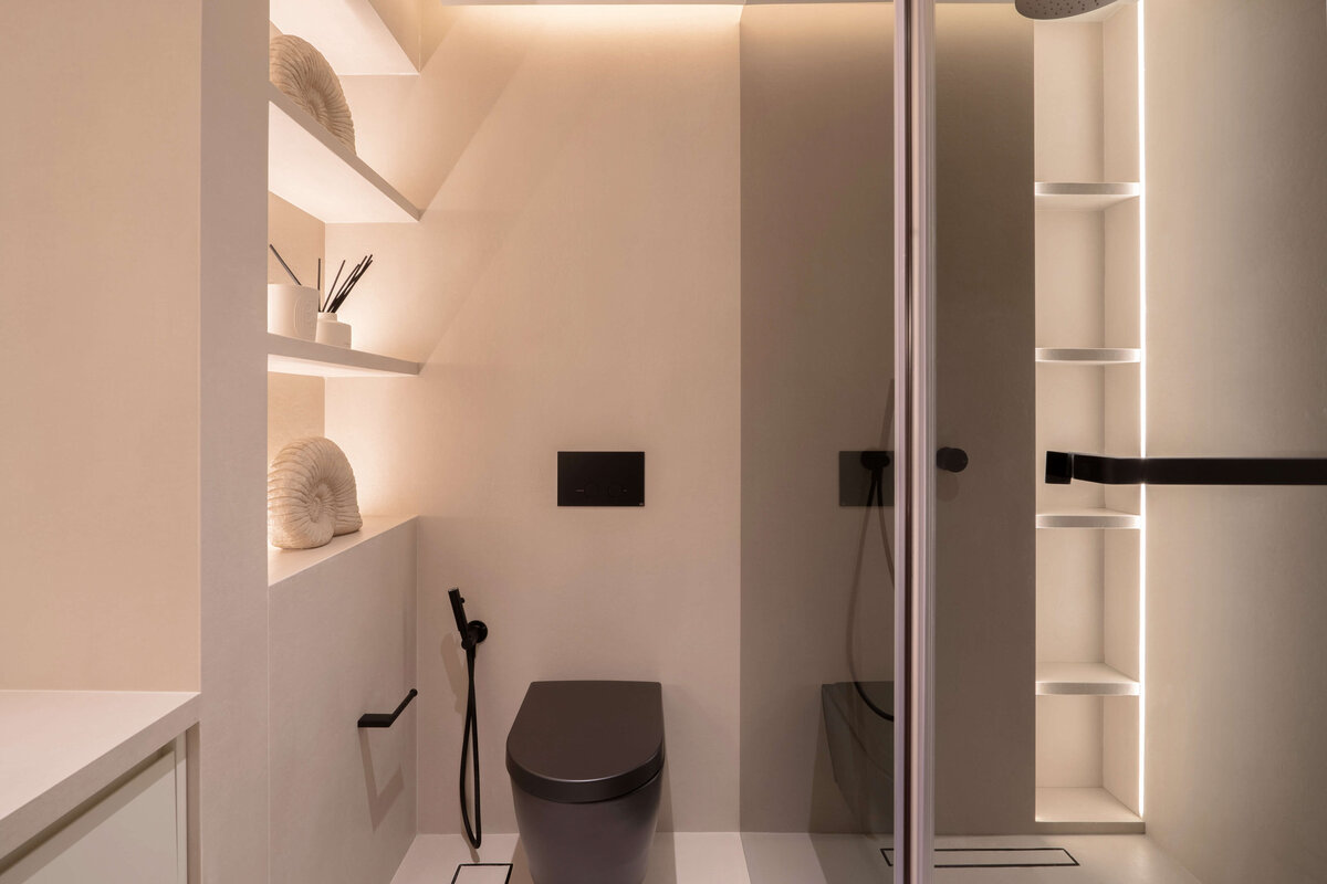 Spacious bathroom with a focus on light, neutral tones. Large built-in shelving emits a soft light. A black toilet adds contemporary flair to the space.