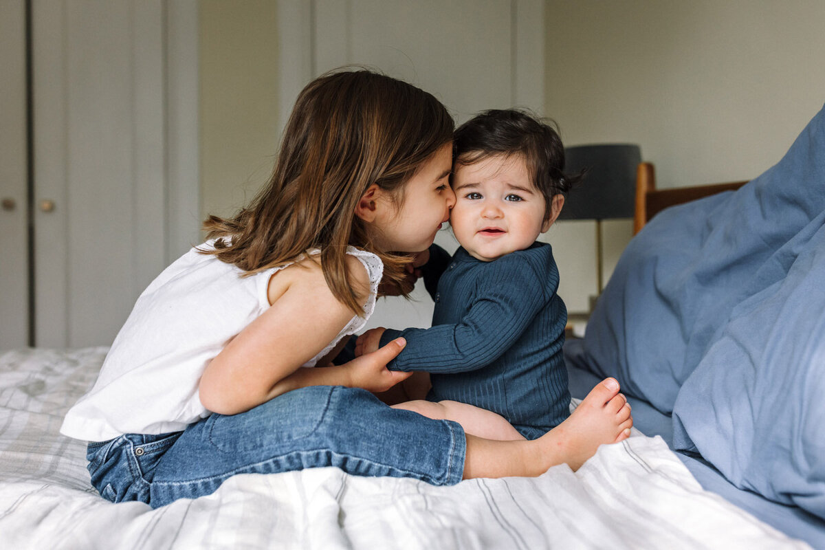 baby sitting on bed looking at camera with older sister leaning her face into baby and holding hands