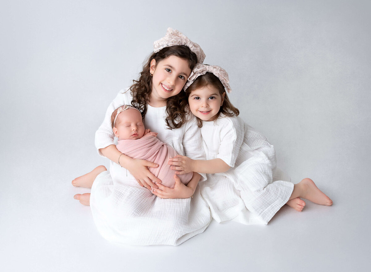 siblings photos  2 older sisters wearing white dresses holding their baby sister wrapped in pink during a professional baby photography session in NYC