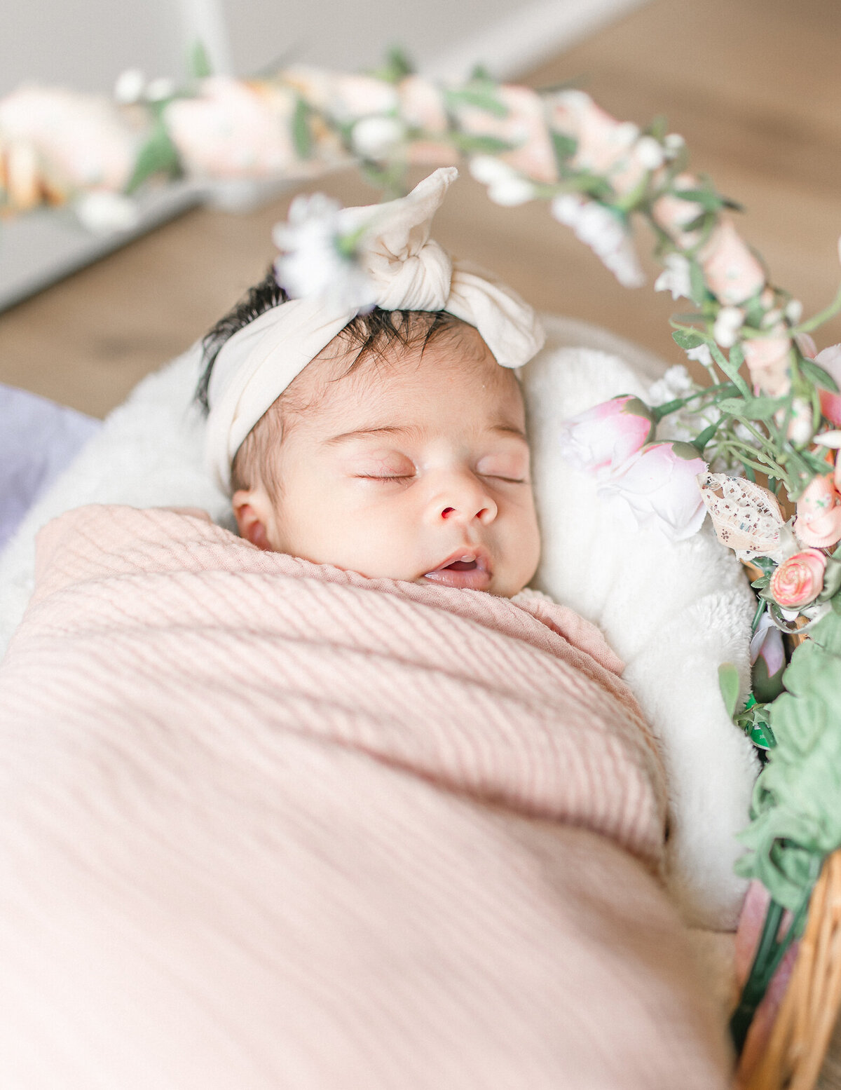 A newborn baby girl sleeps peacefully in a floral-adorned basket, wrapped in a soft blush swaddle with a delicate bow headband.