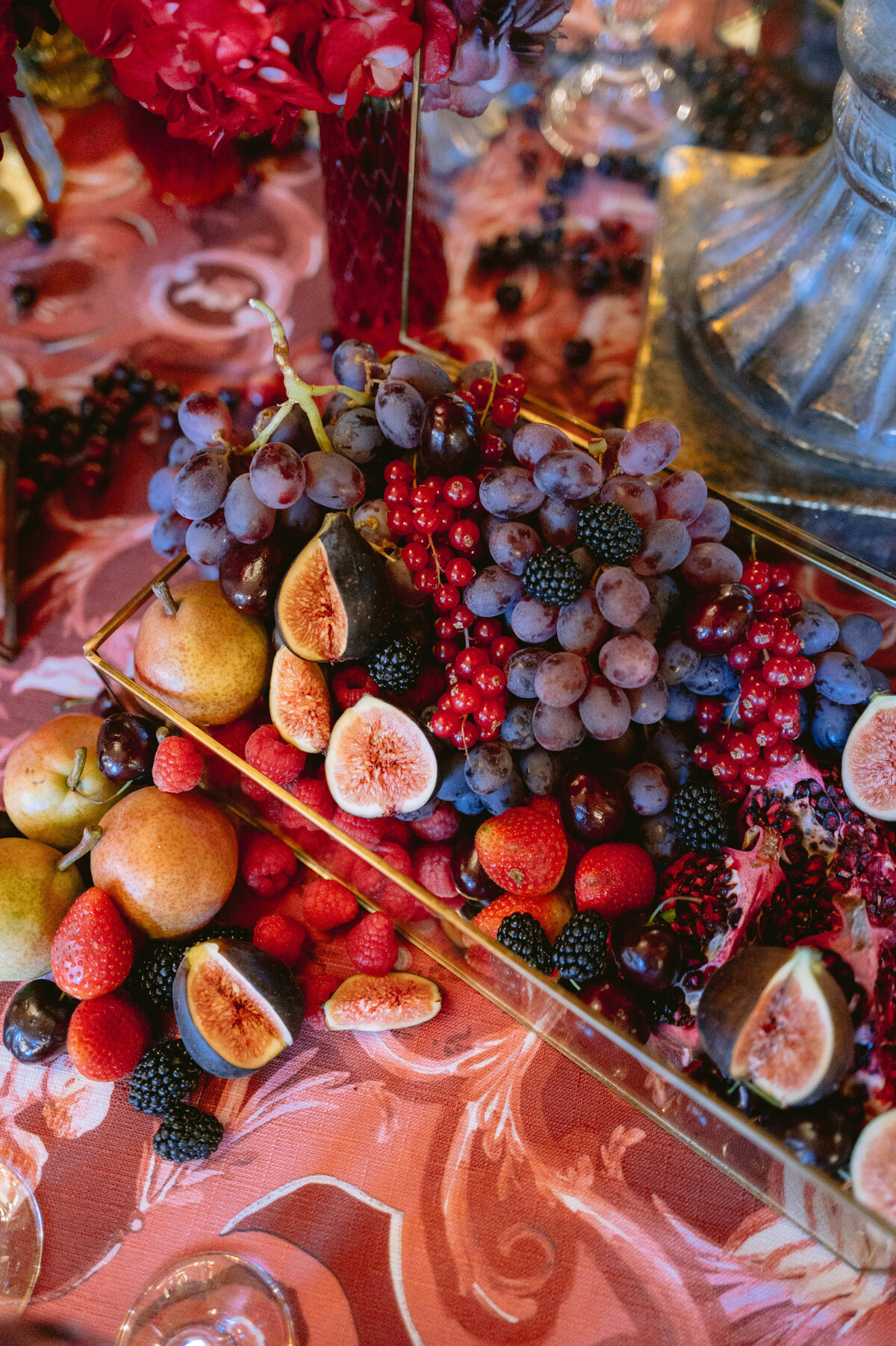 The photo was captured at Shangri La Hotel & Ritz Paris Hotel in Paris, France, by the talented photographer Matteo Coltro, with Zen Film Works as videographers. In the foreground, a vibrant fruit display greets the eye, showcasing clusters of grapes, figs, blackberries, red currants, raspberries, and pears meticulously arranged on an exquisite table adorned with a decorative pink and gold tablecloth. This rich assortment of fruits not only adds a burst of color but also introduces various textures that contribute to the overall festive ambiance of the scene.