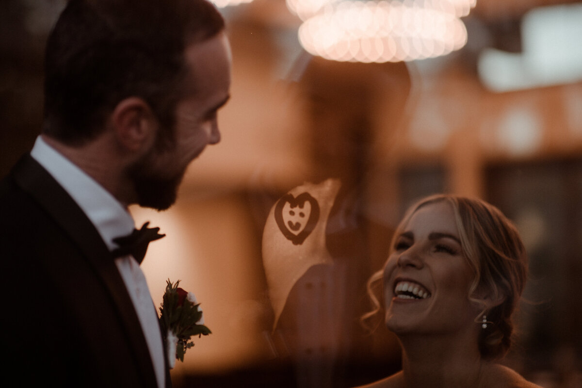 Bride and groom looking at each other with a smiley face inside a heart is draw in condensation on the glass