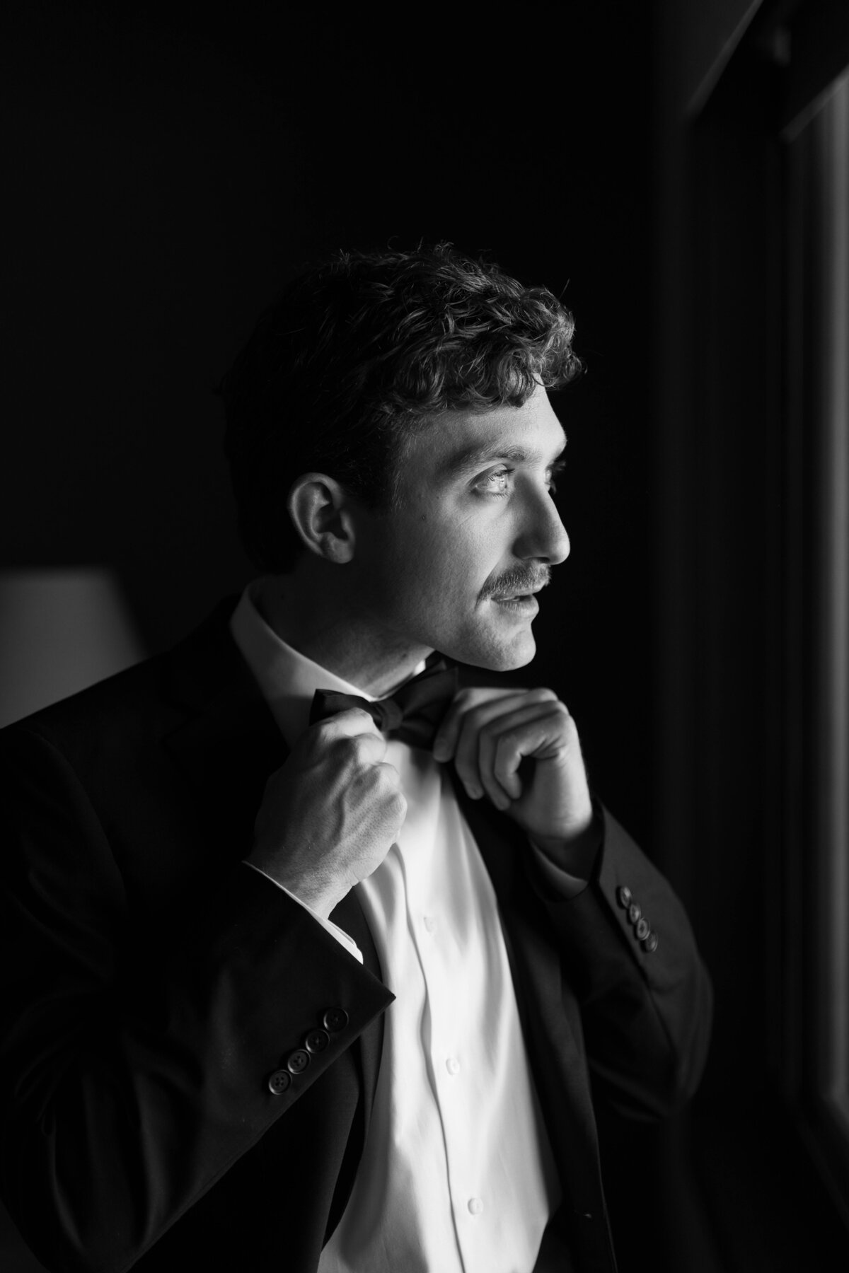 Groom adjusting bow tie while standing near window in black and white