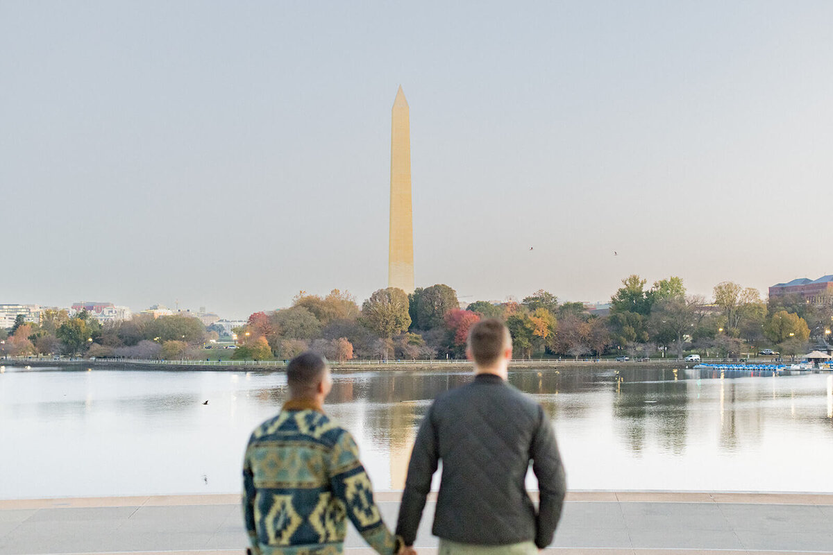 Romantic engagement photo of a same-sex couple holding hands and gazing at the Washington Monument in Washington, D.C. during golden hour. The Tidal Basin reflects the soft evening light, with autumn foliage adding warmth to the scene. Captured by an LGBTQ+ wedding photographer specializing in authentic and inclusive engagement photography.
