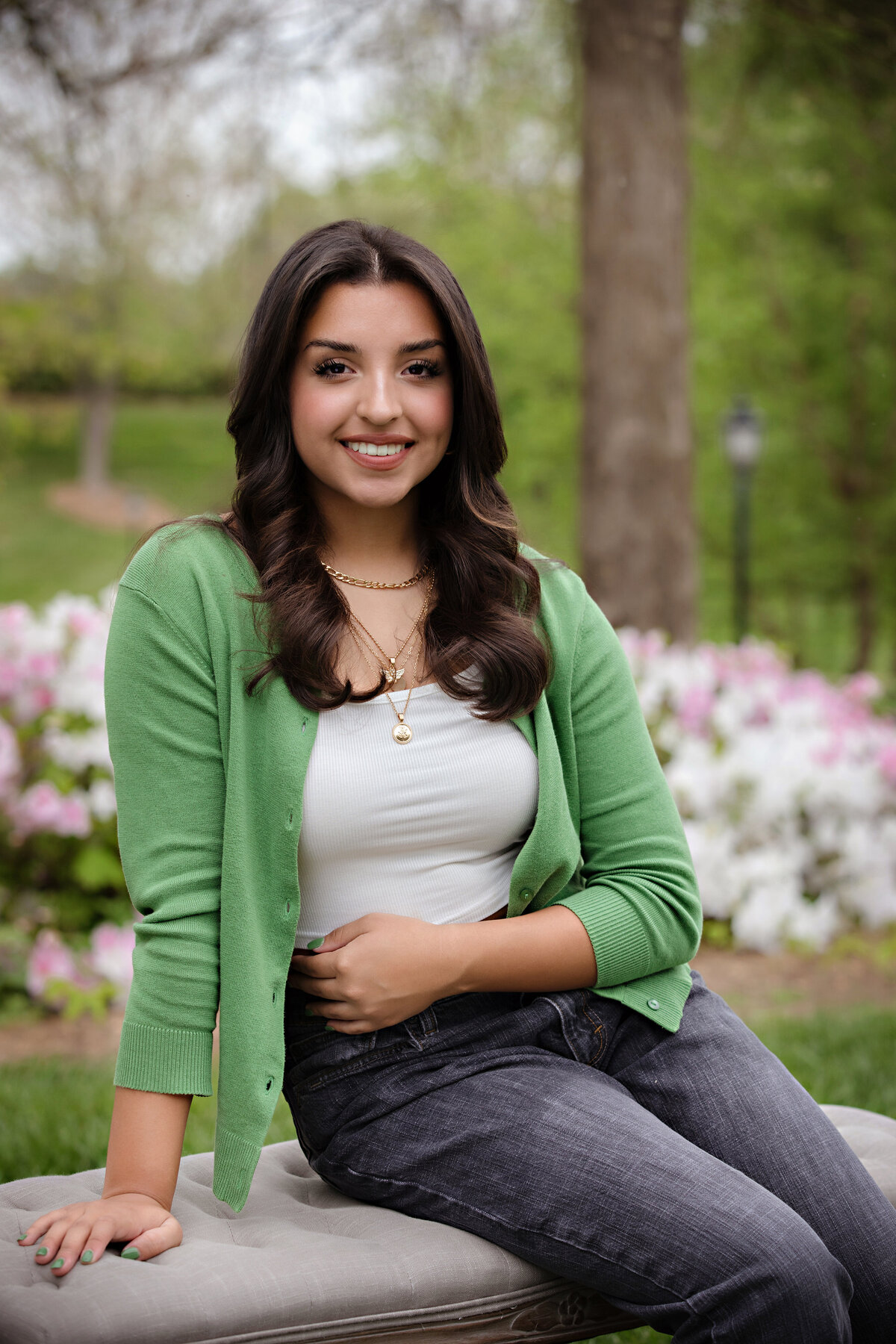 High school senior sitting on a bench surrounded by blooming white and pink azaleas in spring, wearing a green cardigan, white tank top, and gray jeans.