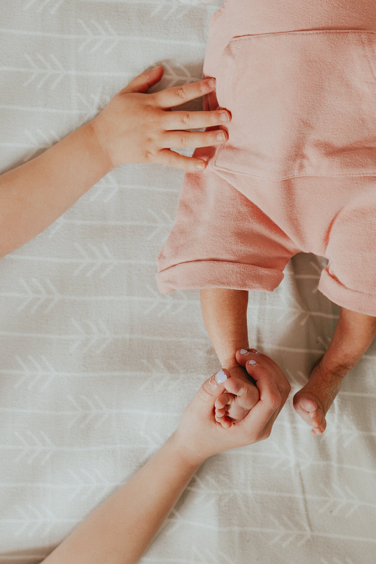 Photo of a newborn baby laying in her crip where you can just see her legs and her siblings hands holding onto her taken by a Langley Family Photographer