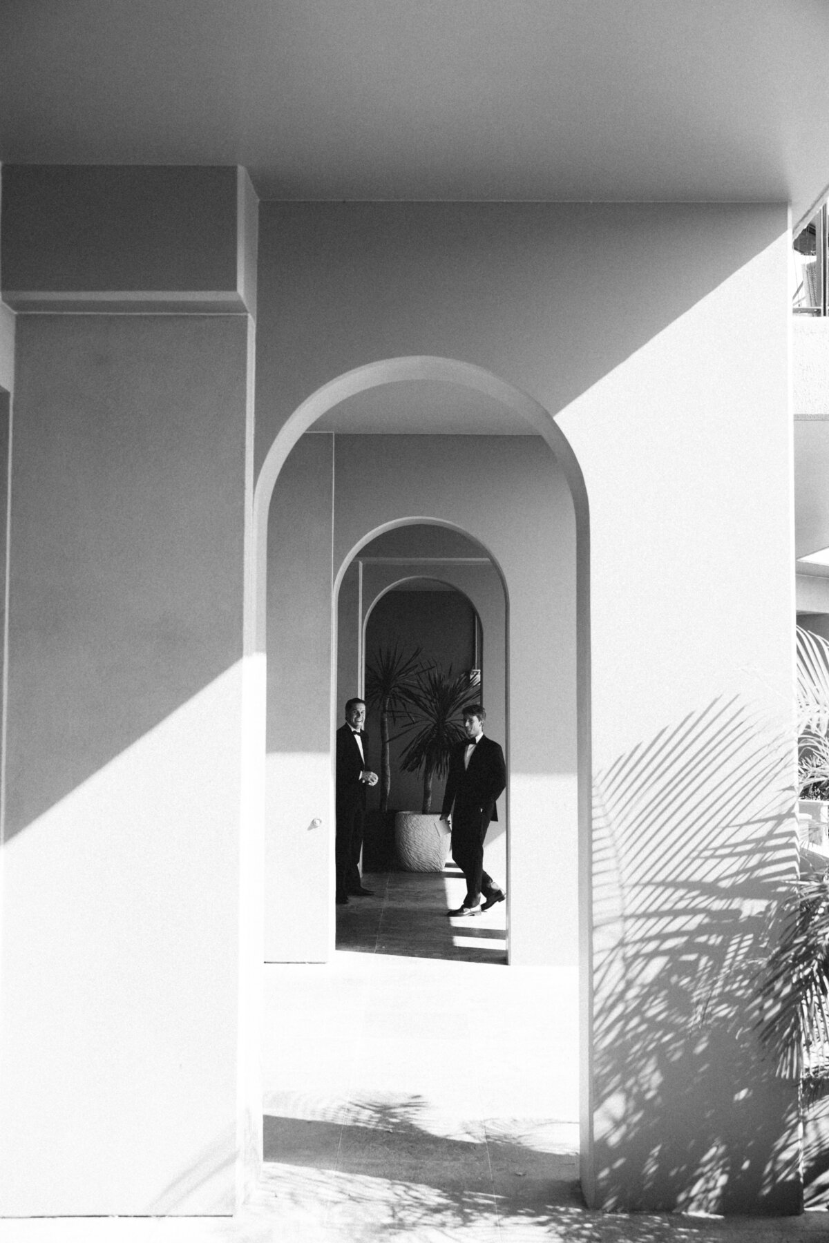 Two groomsmen in tuxedos stand under modern archways at 1 Hotel Hanalei Bay, with dramatic light and shadow creating an architectural composition.