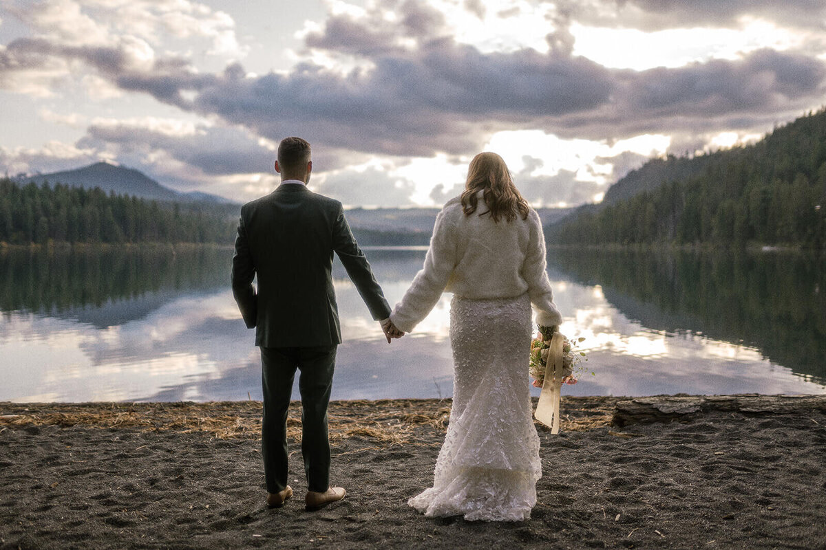 A bride and groom hold hands on the lake shore facing away from the camera.
