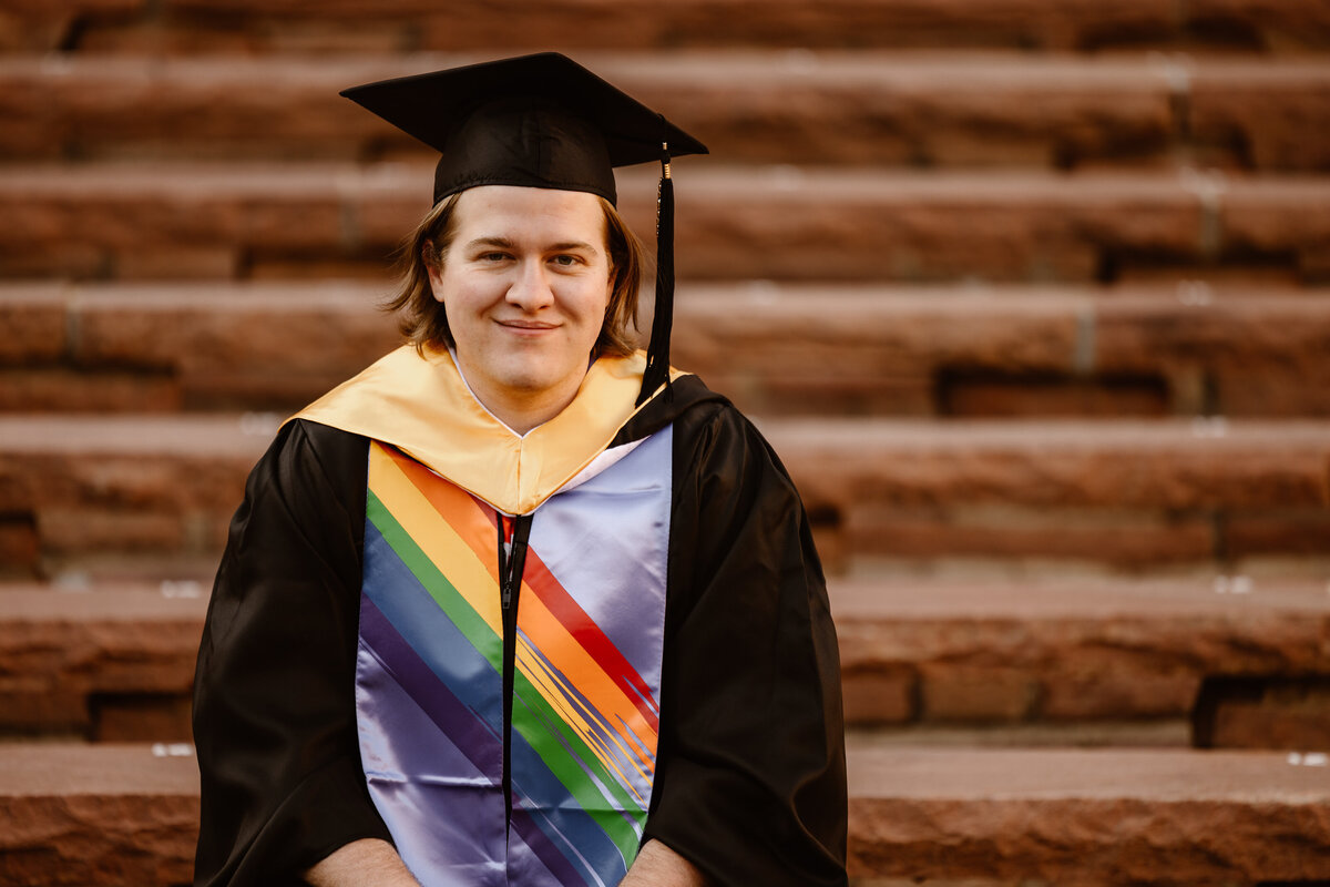 A graduate in cap and gown smiles while seated on the steps at CU outdoor theater, proudly wearing a colorful pride stole.