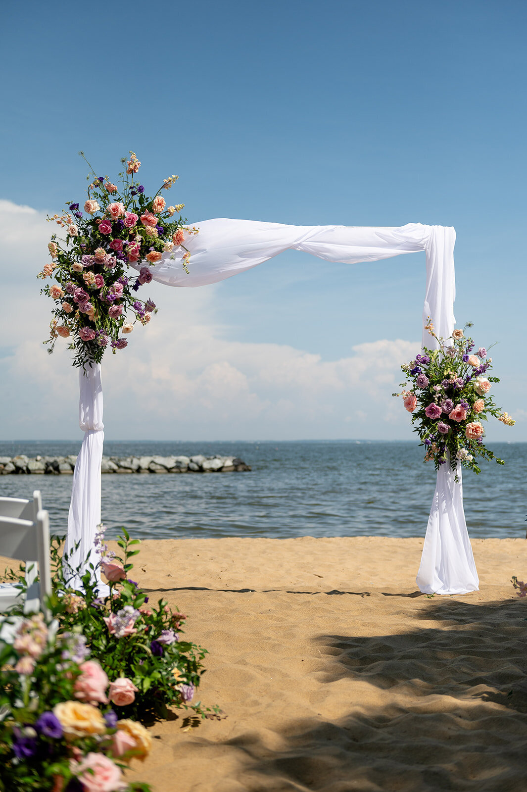 Ceremony arch elegantly draped with white sheer linens and adorned with white and blush florals and greenery, set on a sandy beach with a waterfront view of the beautiful Chesapeake Bay. Foundation.