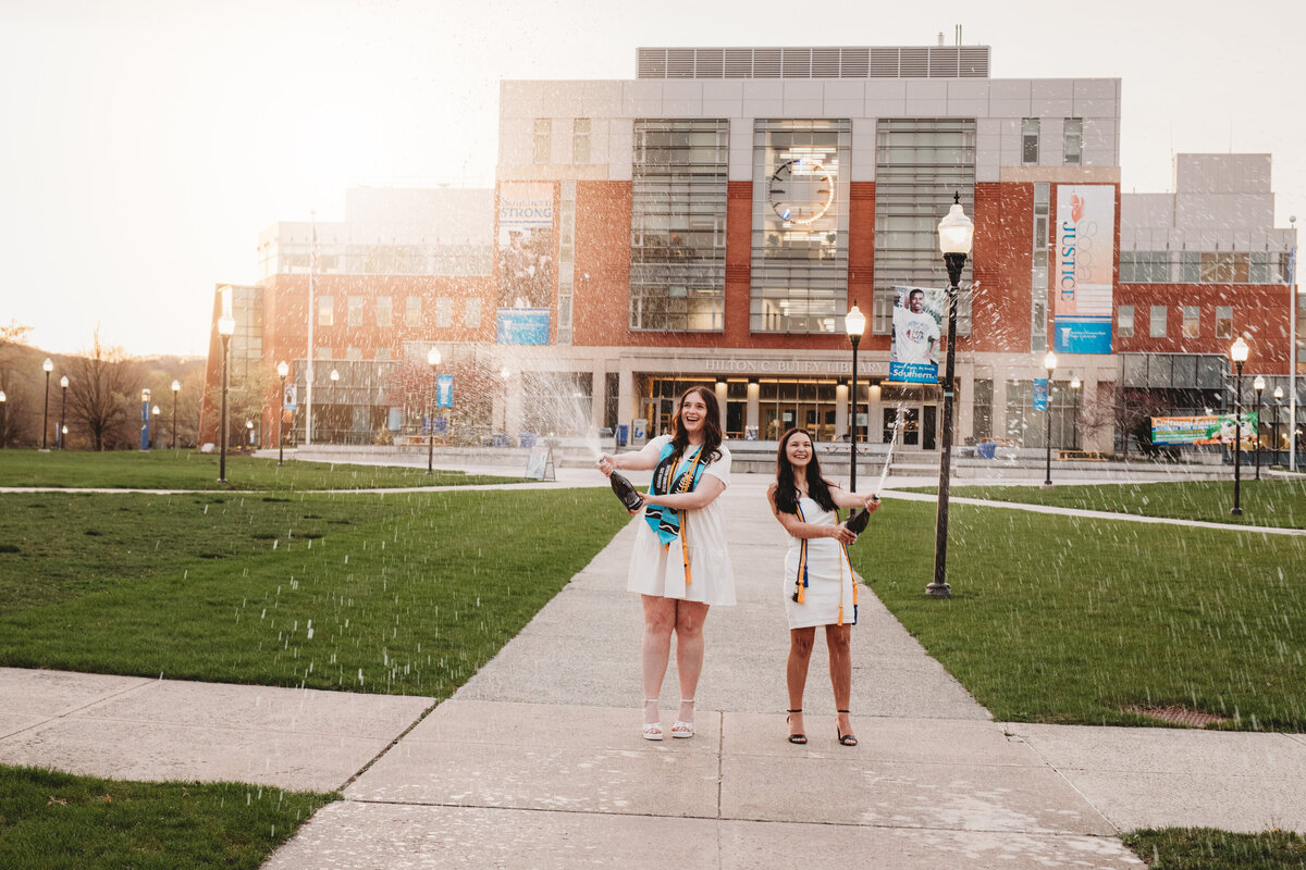 Two Southern Connecticut State University graduates in white dresses and gold stoles celebrate by popping champagne in front of the university building at sunset. Captured during a Connecticut college graduation photography session by Nicole Mele Photography, showcasing excitement and achievement.