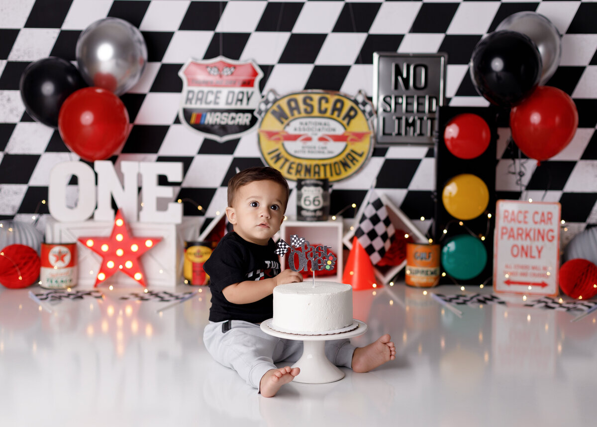 Racecar Nascar themed cake smash at West Palm Beach, FL newborn photography studio.  Baby boy sitting behind untouched white cake smiling at the camera. In the background, there is a black and white checkered backdrop, black, white, and red balloons, a stop light, and race flags.