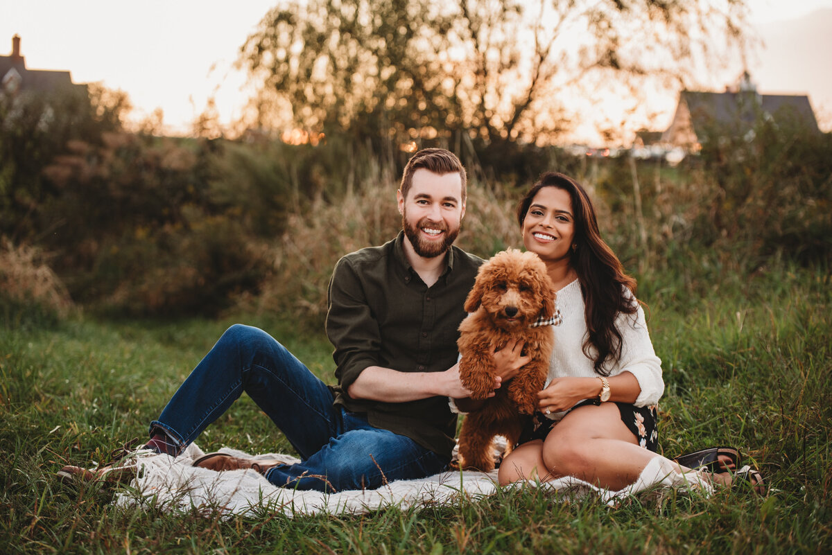 A smiling couple sits on a cozy blanket in a grassy field at sunset, holding their adorable fluffy dog during a lifestyle photography session in Connecticut. Captured by Nicole Mele Photography, celebrating love, companionship, and furry family members.