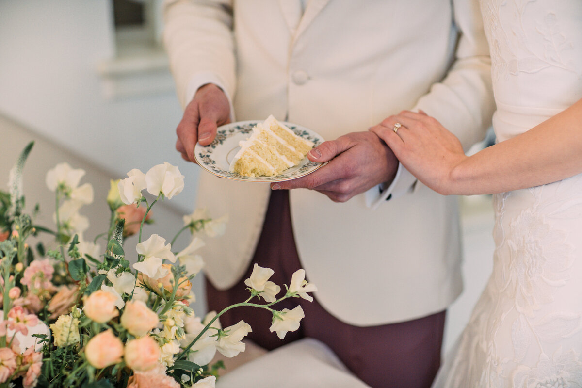 Closeup of a slice of Zingerman's Bakehouse buttermilk wedding cake on an antique plate held by a bride and groom near flowers