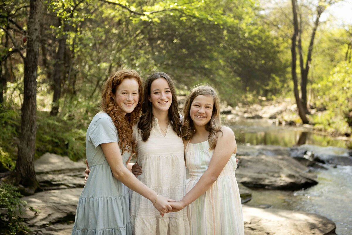 Three sisters happily standing together by the creek in Flower Mound, their arms around each other as they smile for the camera