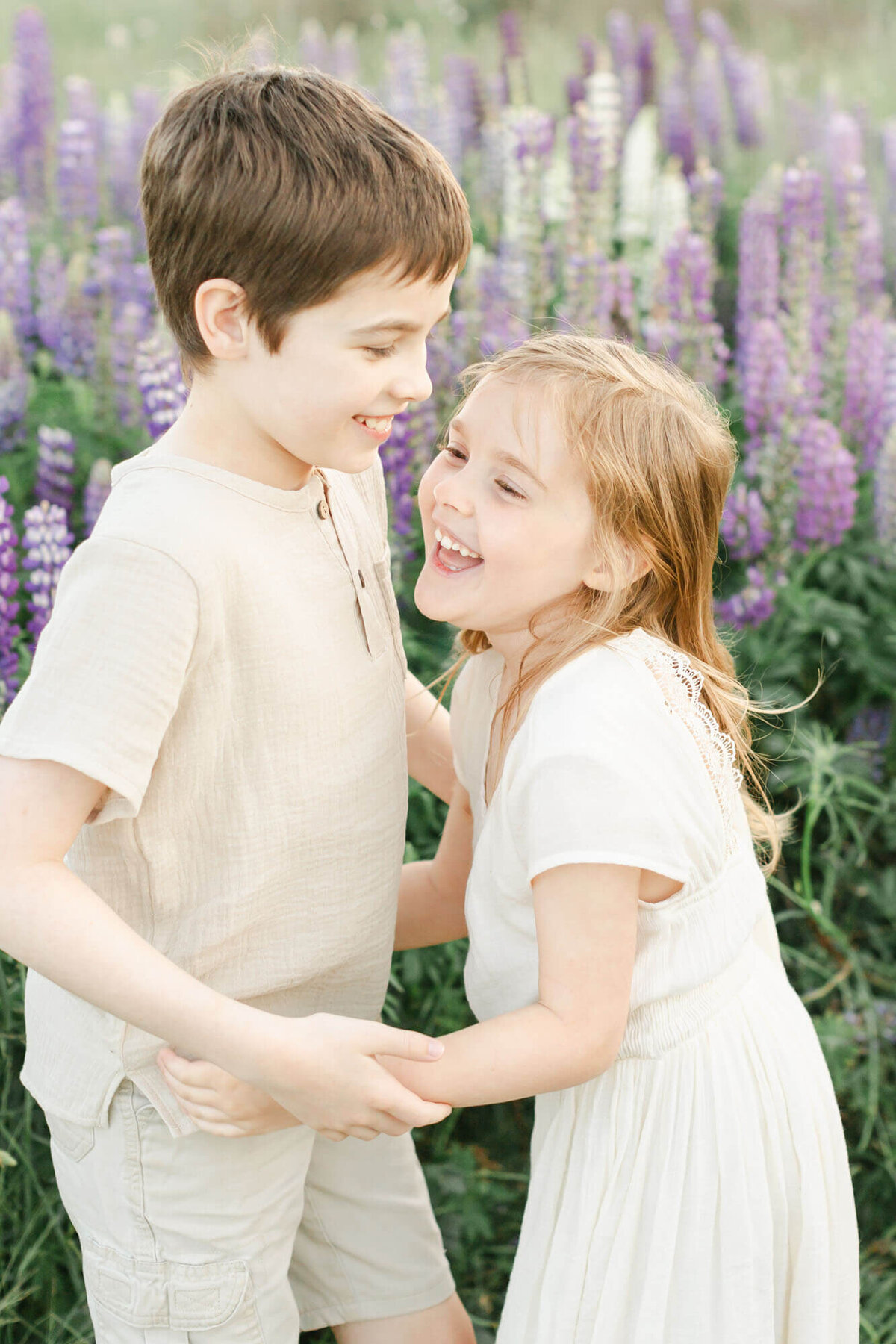 Boy who is about 8 is holding his sisters arms, sister is about 5. They are laughing with one another and standing in a field of purple and white lupine wildflowers. They are both dressed in light cream and taupe.