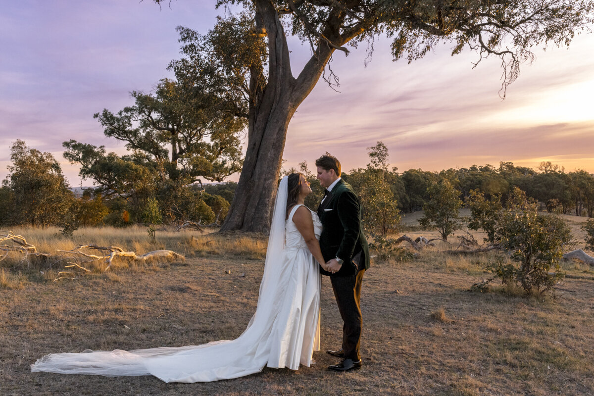 Bride and Groom standing on a mountain top. The wind is blowing in her hair and they are looking out to the view while hugging each other. The bride is a long sleeved wedding gown.