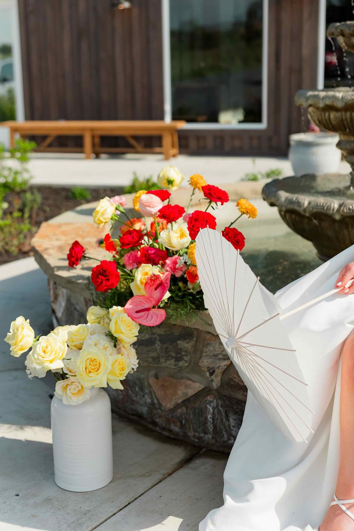 Outdoor wedding ceremony decor with bright florals in white vases beside a fountain.