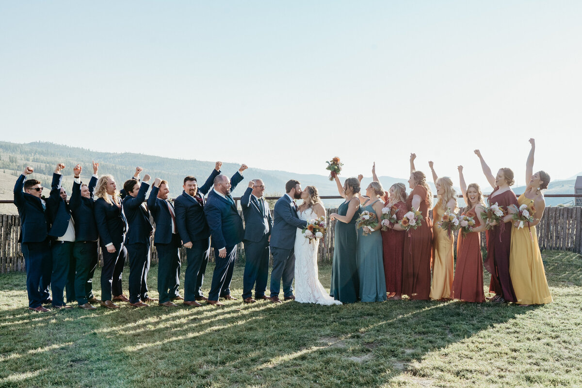 Bride and groom pose with their wedding party including bridesmaids in multi colored dresses.