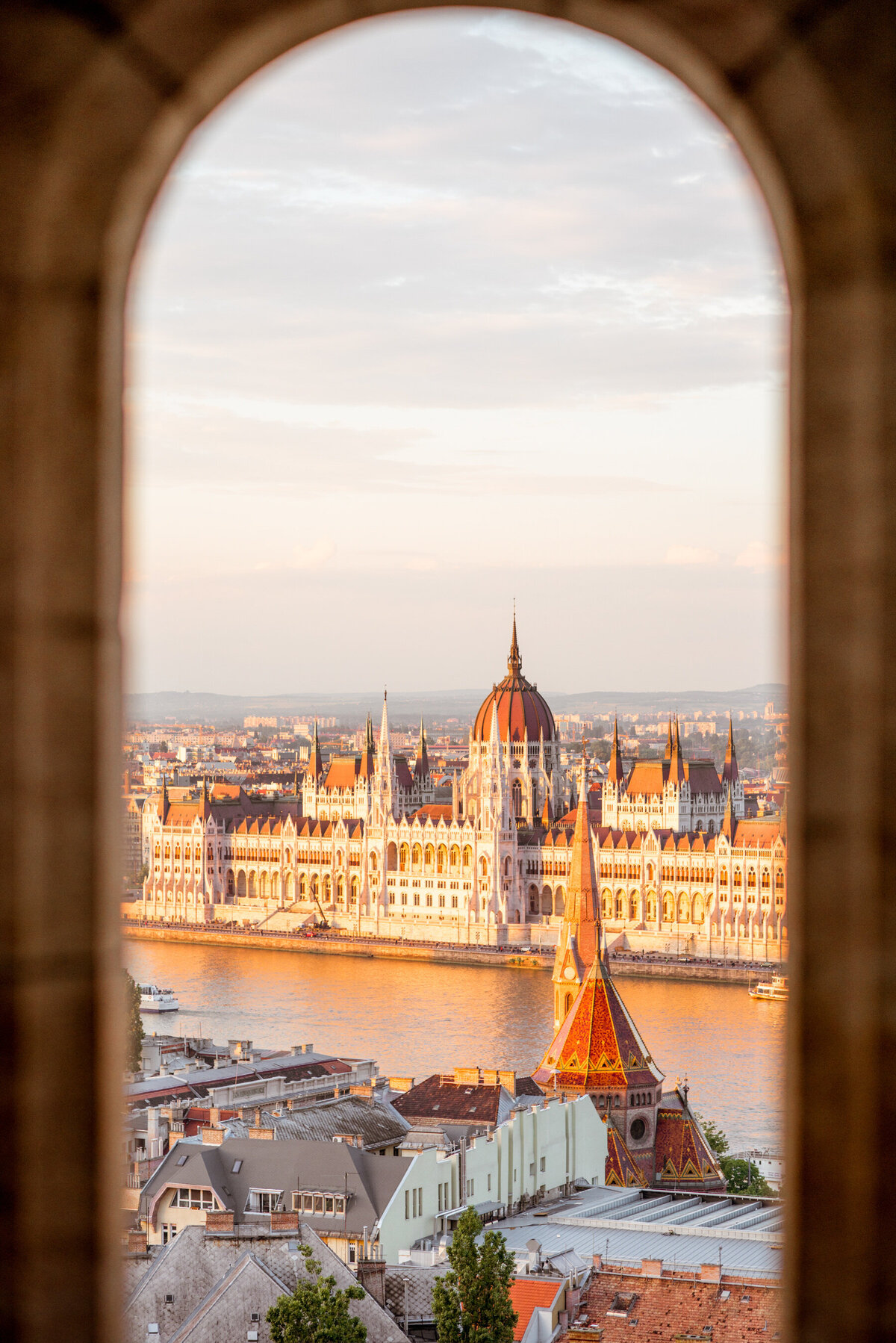 View through an arched window of the Hungarian Parliament Building in Budapest, bathed in golden light. The Danube River and rooftops of nearby buildings evoke the charm of Caribbean getaways, creating a picturesque urban scene.