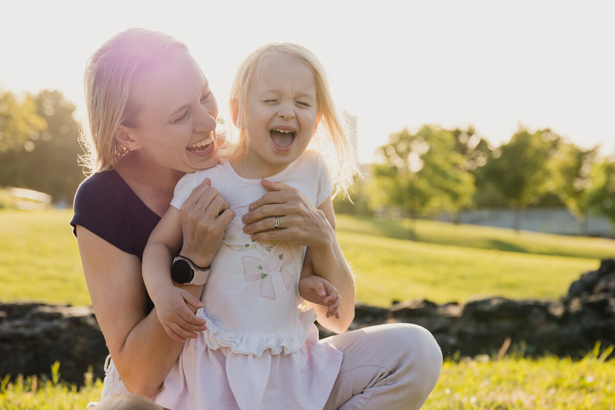 A woman laughing and holding a young girl in a park, both smiling brightly in the sunlight.