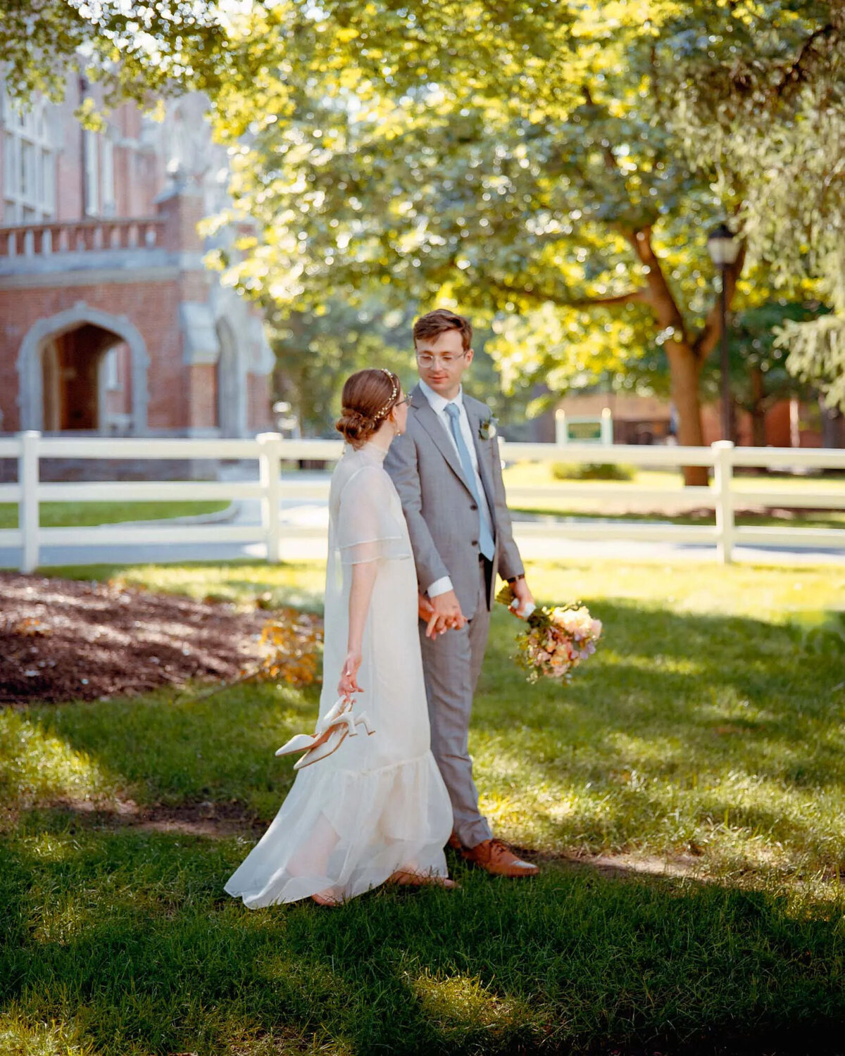 A bride and groom stand hand in hand on a sunny lawn, captured by a film photographer NJ. The bride holds her shoes and bouquet, her white dress glowing beside the groom’s gray suit. Trees, white fence, and a brick building frame the moment.