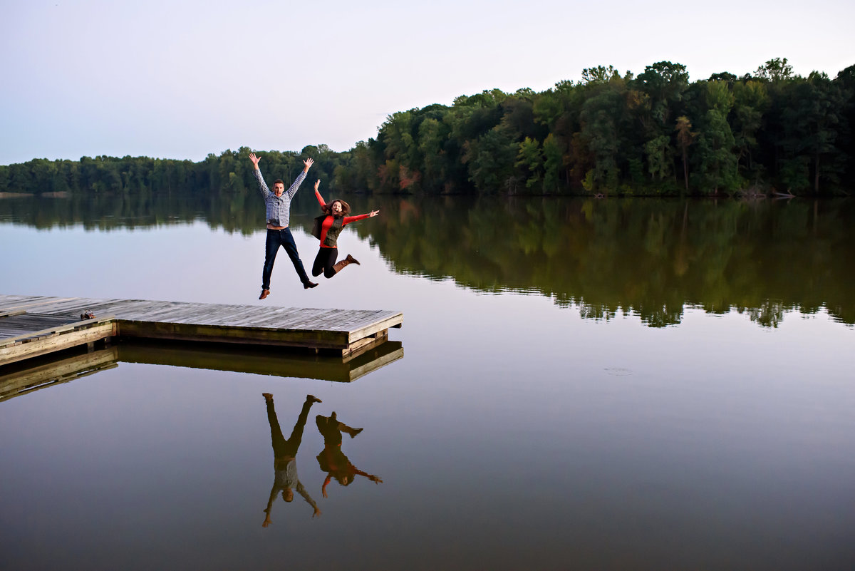 An engaged couple jump at the end of a dock on a lake.