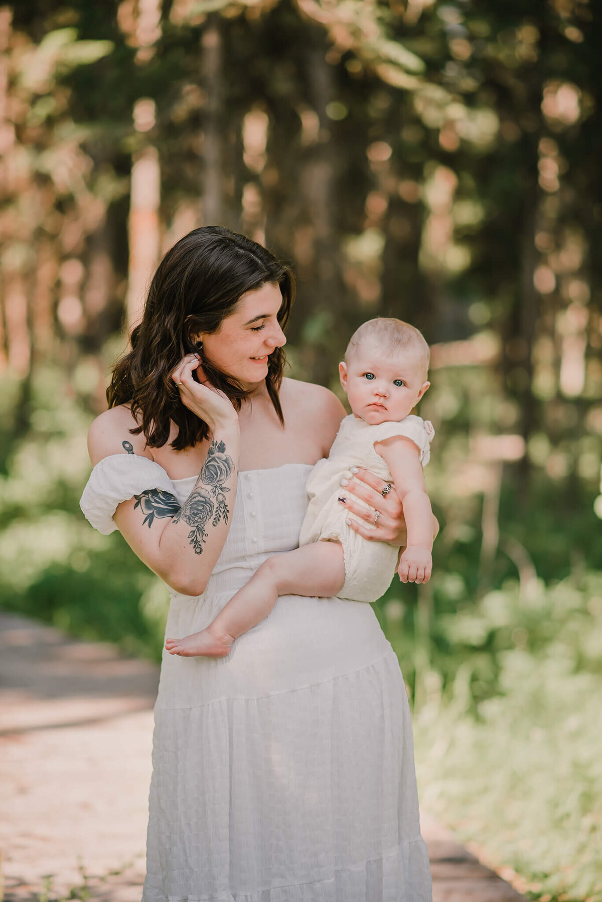 A mother with short brown hair wearing a white dress hold her infant daughter on her hip while looking at her and smilling. Portrait by Brittany Danielle