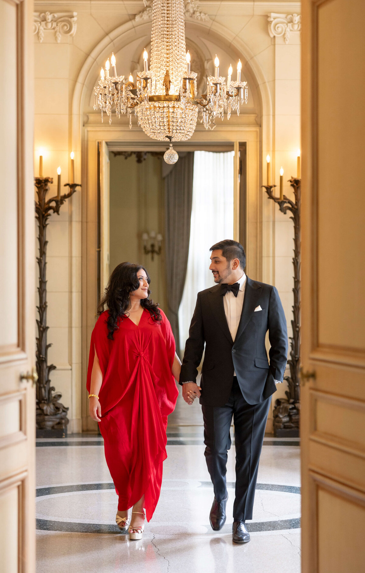A couple elegantly walks hand in hand in a grand, chandelier-lit hall. The woman wears a flowing red dress, and the man is in a black suit with a bow tie. They are smiling at each other, framed by ornate doors.