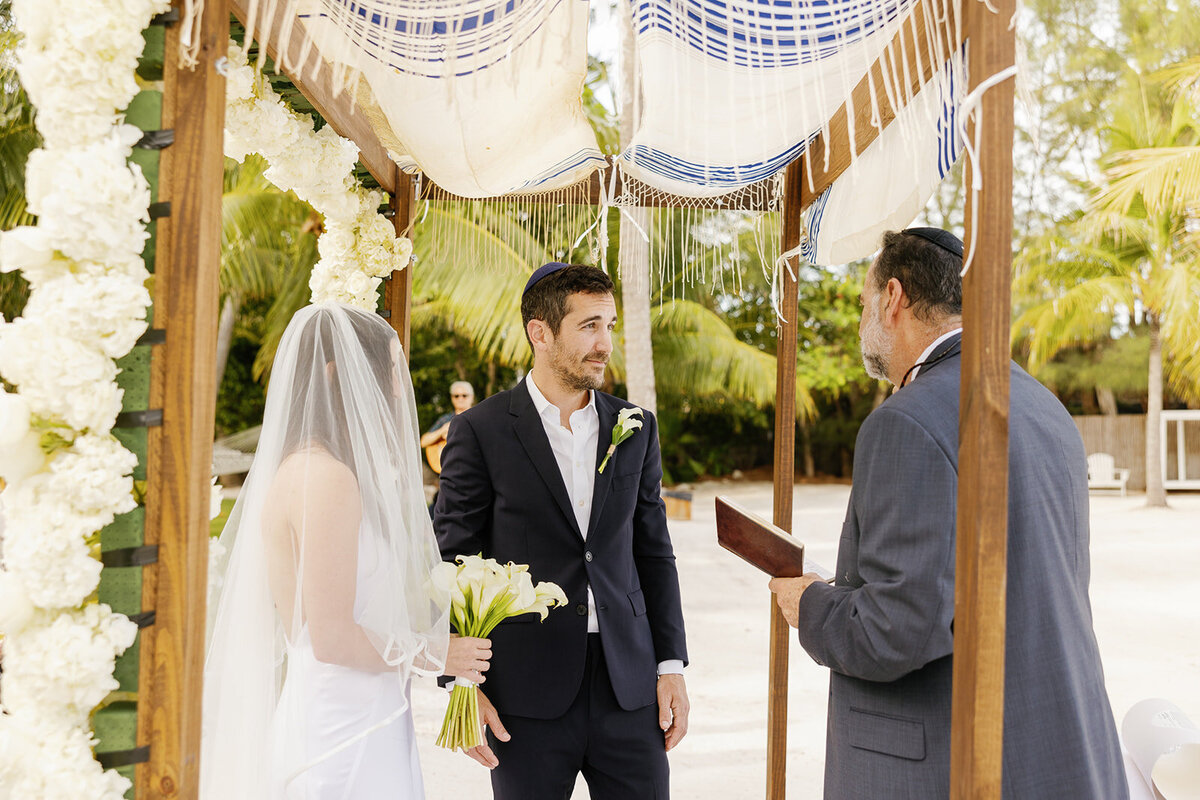 Minette and Evan exchange vows under a floral chuppah with the ocean in the background. Florida Keys micro wedding captured by Claudia Amalia Wedding Photography.