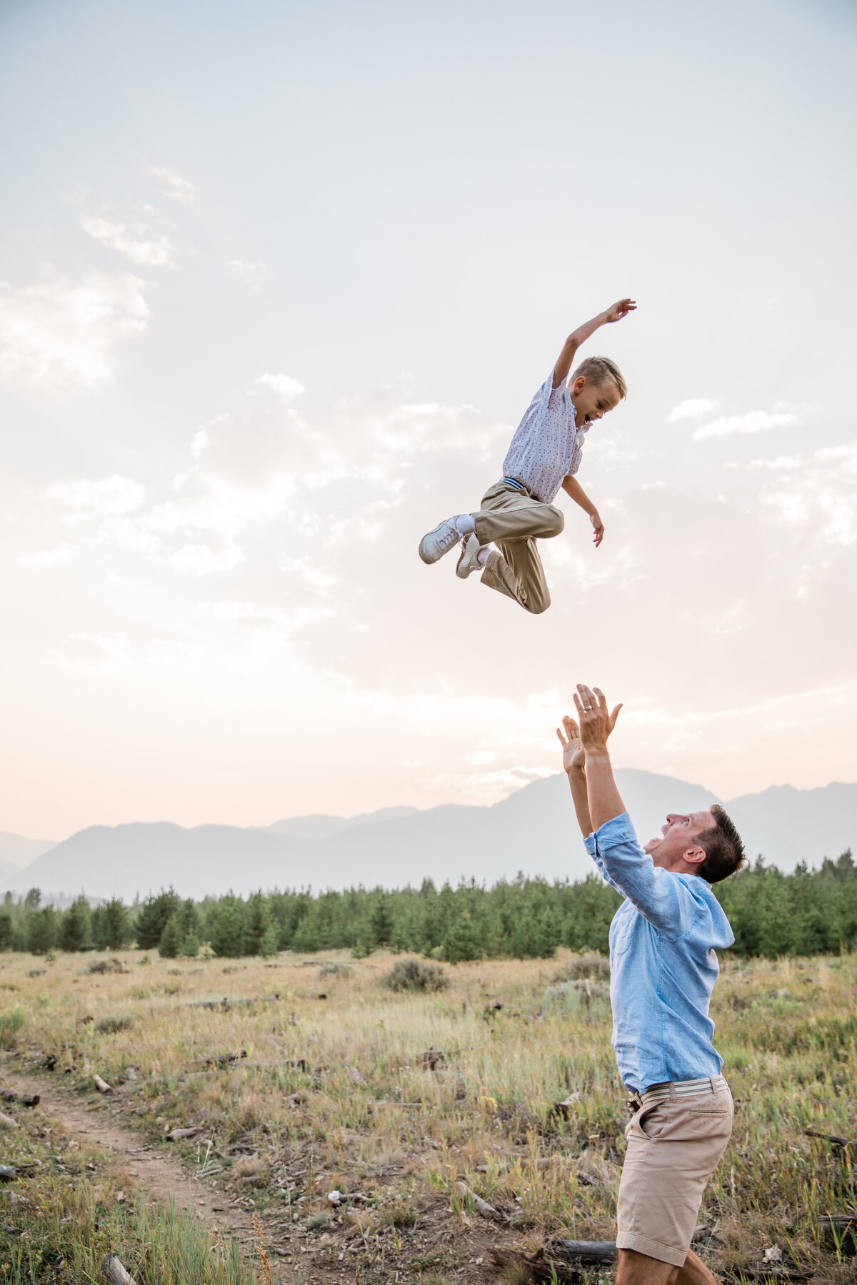 A dad throws his young son playfully in the air. They are smiling with mountains behind them