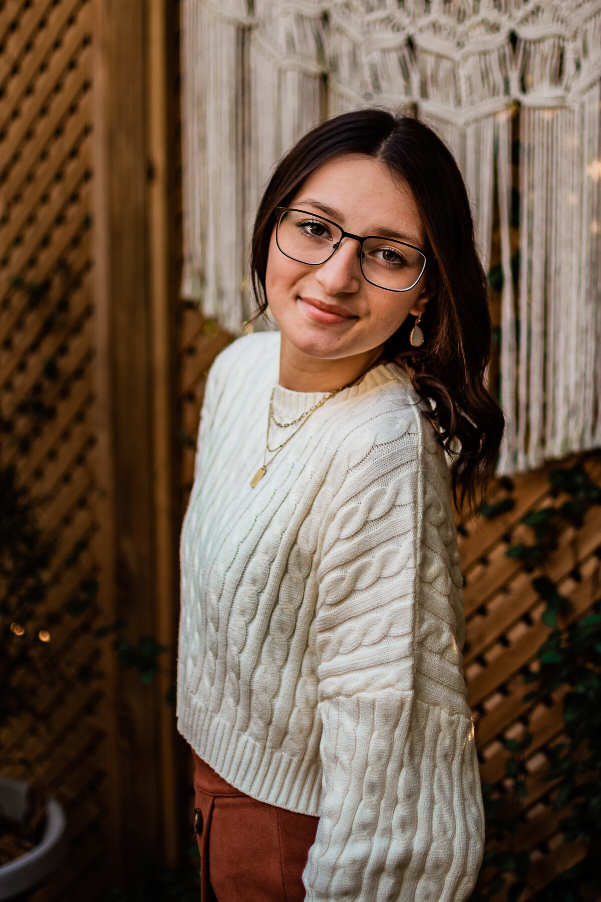 A senior wearing a white sweater stands in front of a boho inspired backdrop and smiles at the camera.