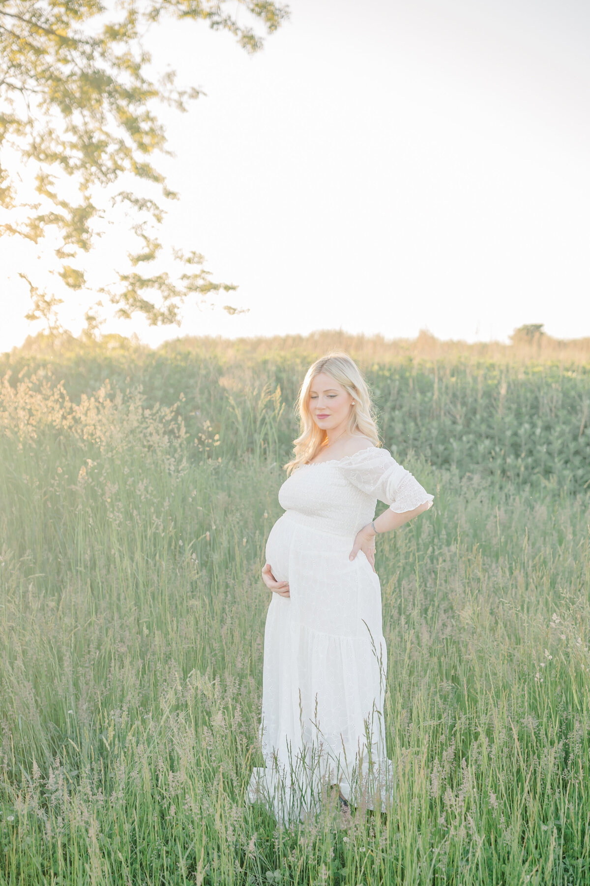 Pregnant mother standing in a lush green field, gently holding her belly, with soft sunlight illuminating her serene expression, captured by a Connecticut maternity photographer