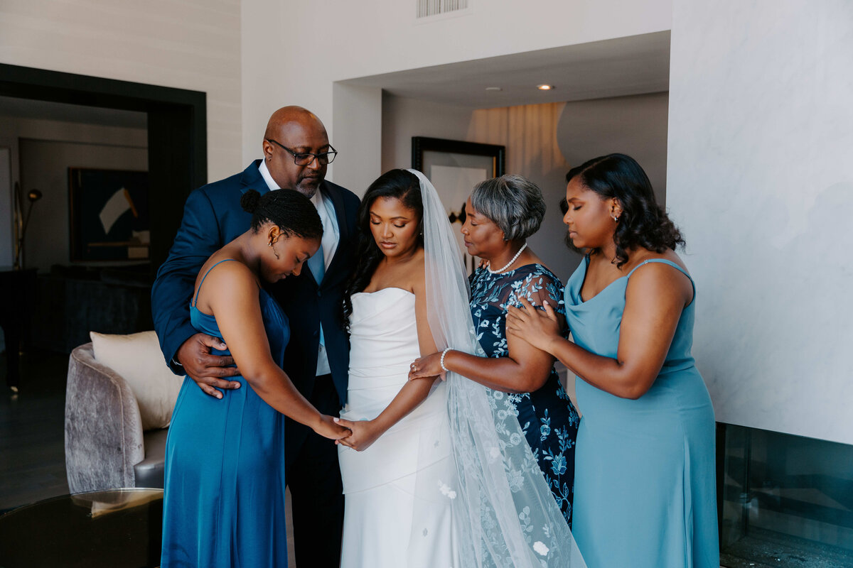 Bride surrounded by family in heartfelt moment of prayer before wedding ceremony.