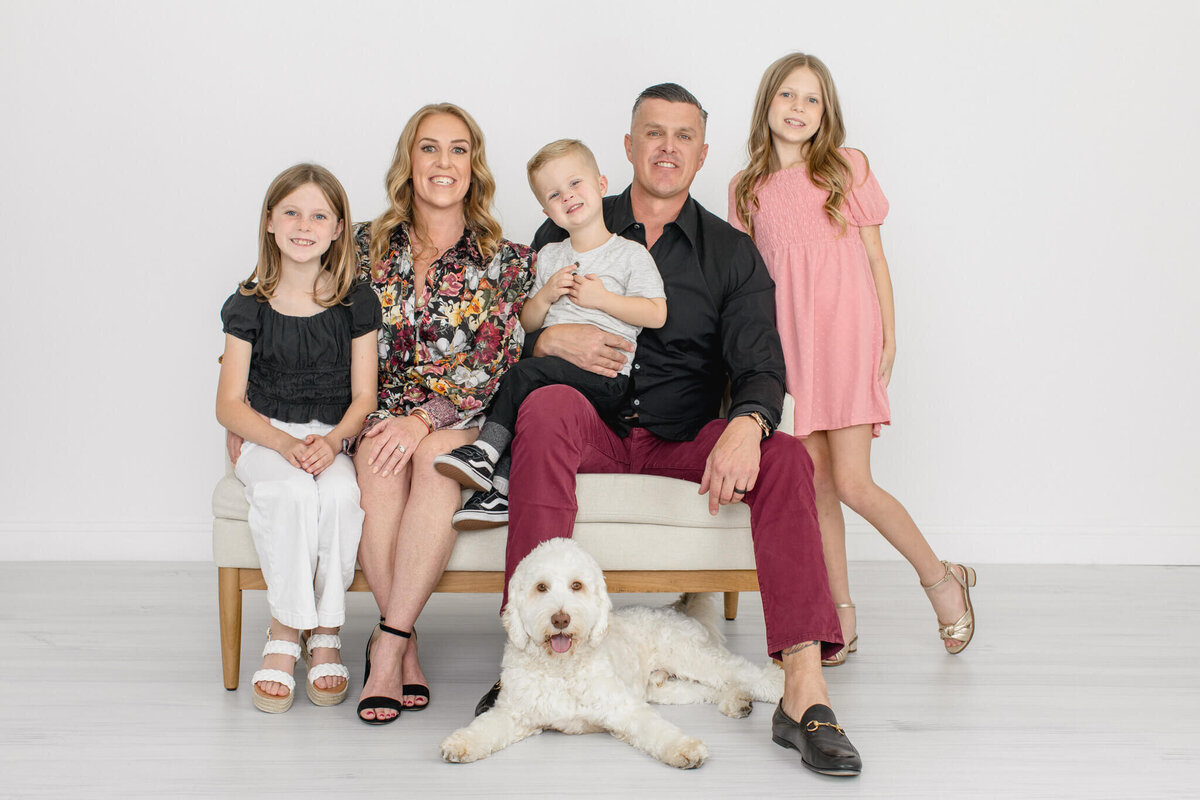 A family of five and their dog sitting on a bench and smiling, wearing shades of pink and black against a white backdrop in a studio in Tustin.
