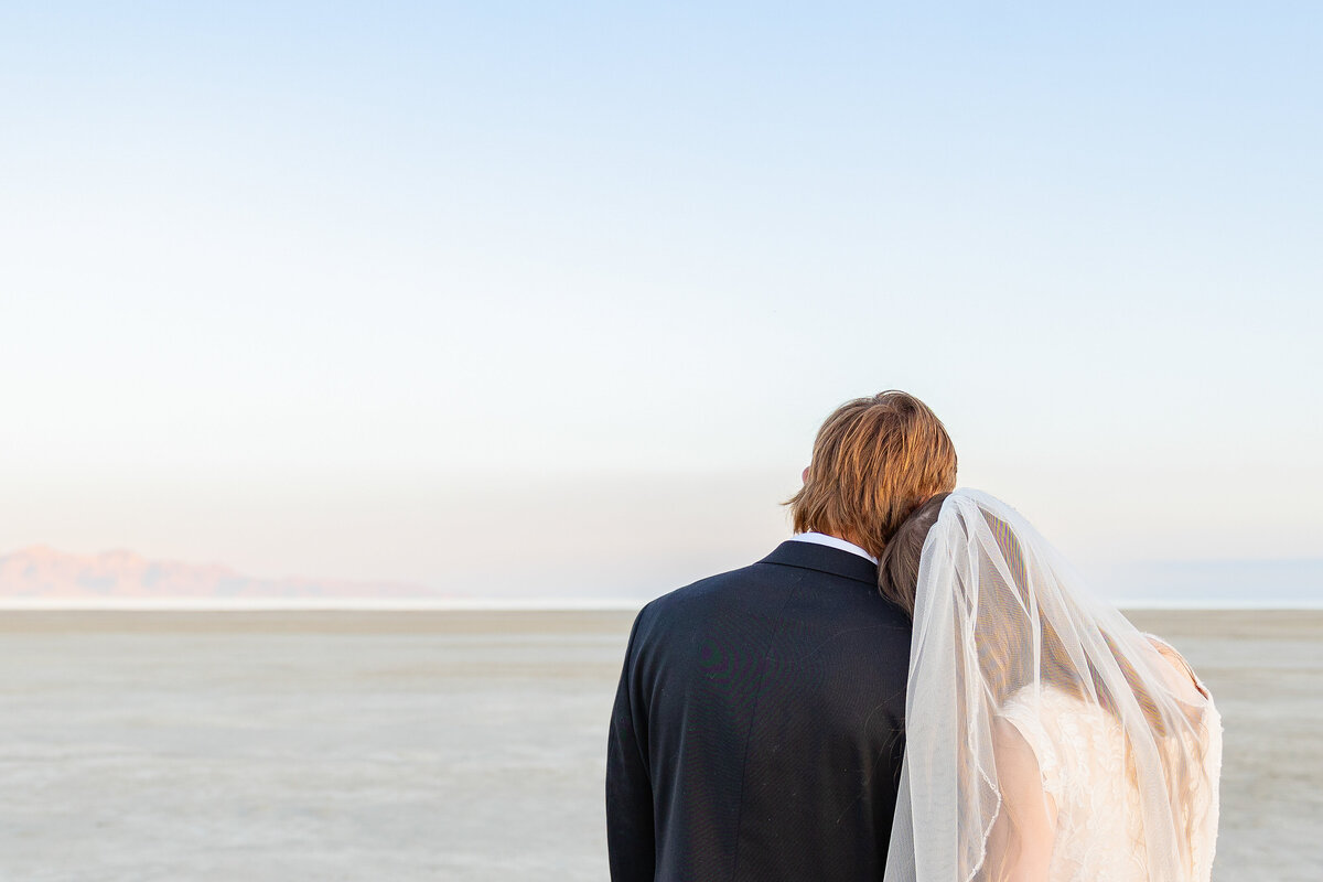 Bride in a flowing veil leans on the groom’s shoulder as they look toward the horizon together.