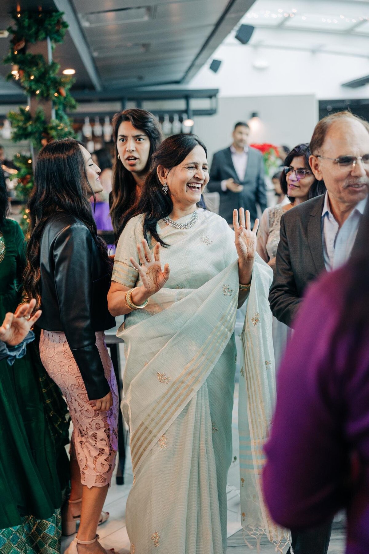 A smiling woman in a sari engaging in conversation at a festive event with other guests in the background.