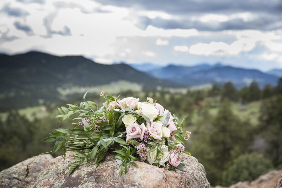 A wedding bouquet with white and pink roses sits on a boulder with a Colorado mountain landscape in the background.