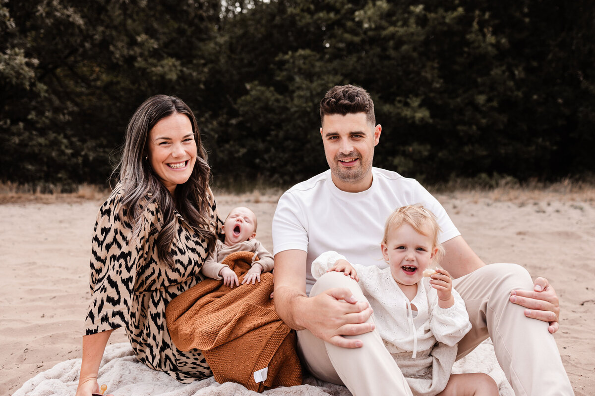 Gezin zit op dekentje dochter zit tussen papa's benen en mama heeft gapende baby op de arm