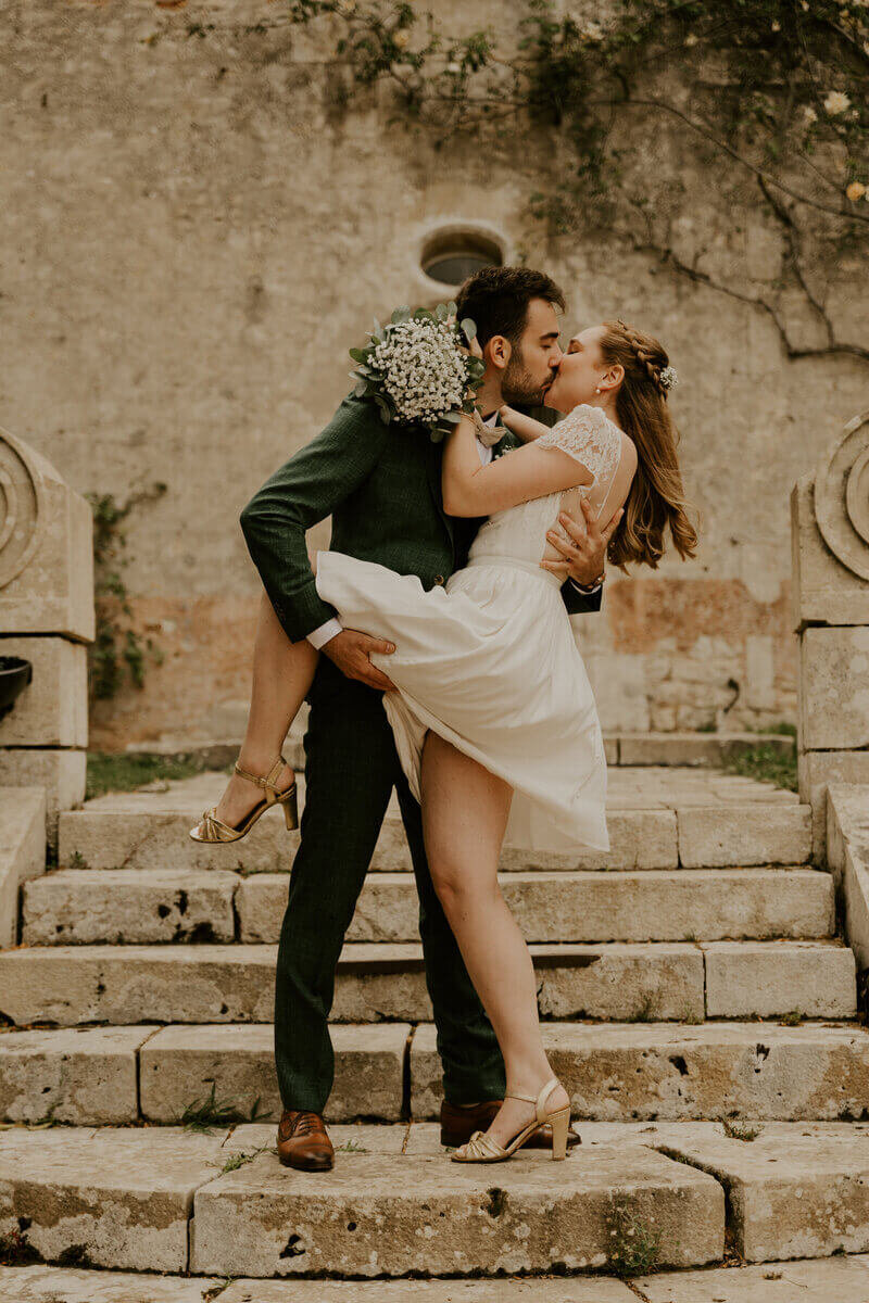 Marié tenant la jambe de la mariée, pendant qu'il l'embrasse et a bascule légèrement en arrière sur des escaliers en pierre. Photo prise par Laura, photographe professionnelle en Vendée.