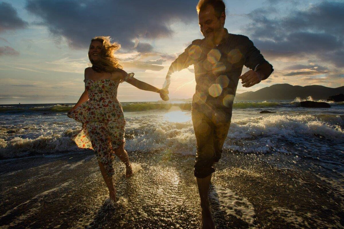 Man and woman run in the ocean water during their engagement session. Water drops are spotted on the lens with the sunset in the background. photo by wedding photographer sacramento, philippe studio pro.